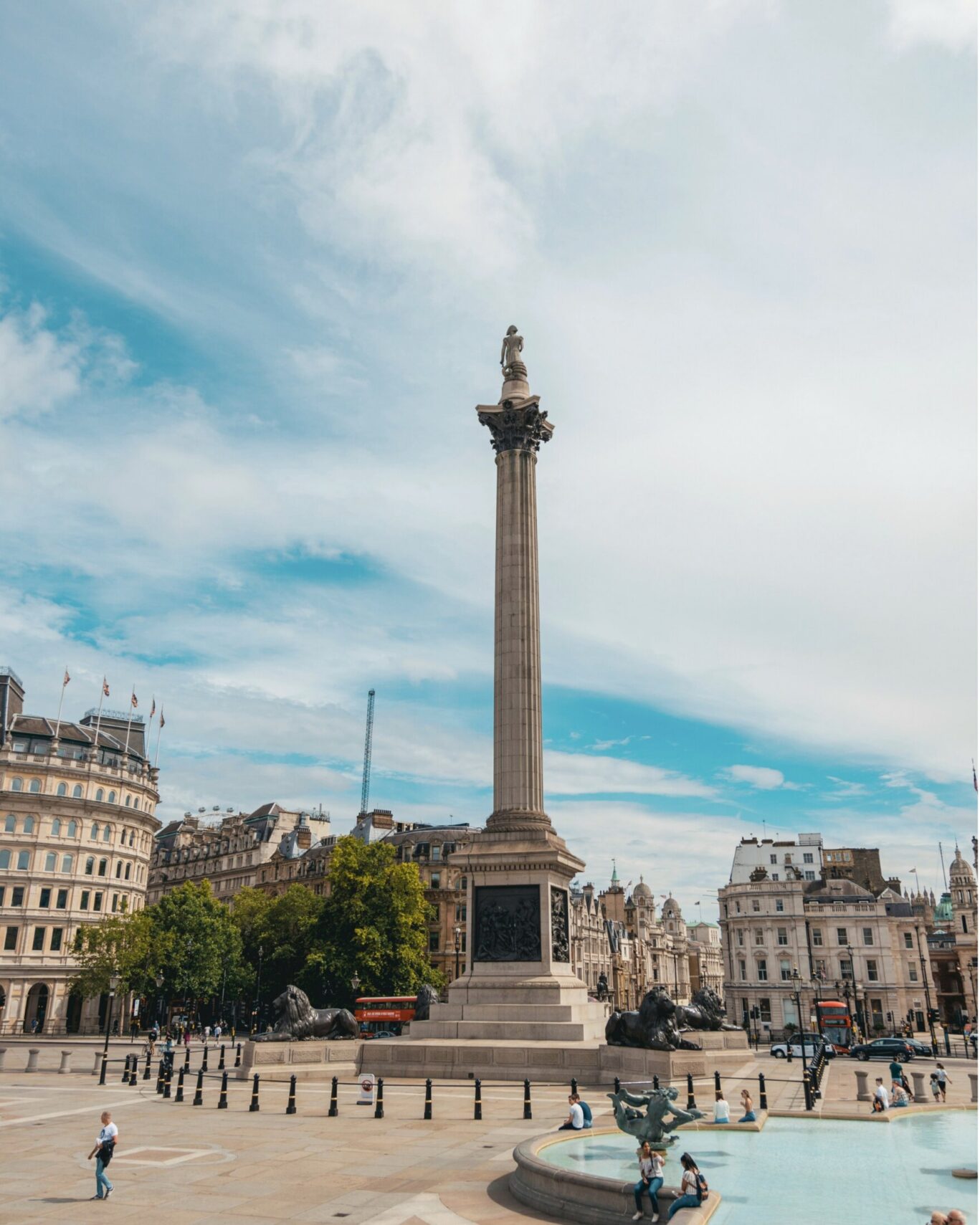 Nelson's Column in Trafalgar Square on a cloudy warm day with several people walking nearby.