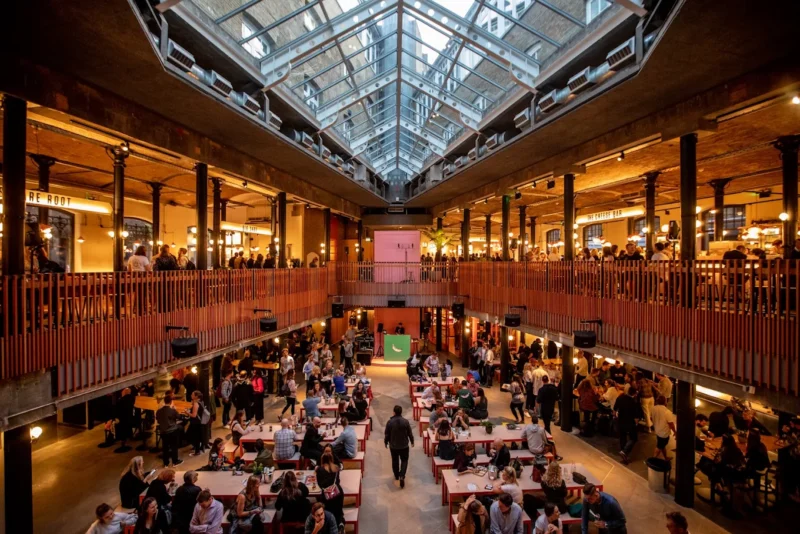 Seven Dials Market, with rows of tables and people sat down with food and drink in a warmly lit food two-tier food court.